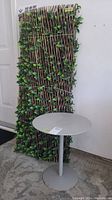Full view of wood and vine craft blind with attached artificial leaves standing upright behind a small round steel table