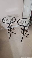 Two steel side tables with round glass tops and nature-inspired metalwork including branches, leaves and small bird figurines, standing on beige carpet in room.