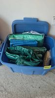 Blue storage tote open showing several folded tarps inside, varying shades of green and blue, with some silver reflective material visible.