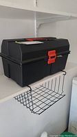 Black plastic toolbox with red latch sitting on white wall shelf.