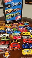 Wide shot of multiple Hot Wheels cars laid out in front of the Hot Wheels carrying case showing case graphics and 38 assorted cars in various colors and styles.