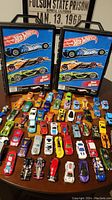 Two black Hot Wheels branded travel cases with handles, and multiple Hot Wheels toy cars in various colors and designs spread around them on a table.