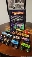 Overview photo of all 15 Hot Wheels Monster Jam monster trucks arranged on a table with the carrying case standing behind them showing Grave Digger artwork on it.
