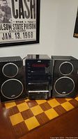 Full stereo system including central unit and two speakers placed on a wooden surface with a black and white music poster in the background.