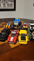 Photo showing nine LEGO vehicles including a large blue and orange monster truck, red and yellow cars in front, and various other colorful LEGO vehicles behind them.