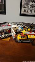Yellow LEGO crane truck with articulated crane arm in front, parked beside semi-tractor truck and RV trailer, plus loose LEGO pieces in bags in the background