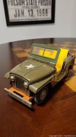 Front-right angled view of the vintage military tin toy jeep showing the green body with white star on hood, front grille, headlights, orange bumper, and yellow seats.