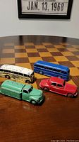 Lot of 4 vintage Dinky Meccano toys displayed on a wood and black checkered board, showing a blue bus, white bus, green truck, and red car.