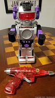Photo showing full view of silver and purple vintage robot toy and red transparent space pistol on a wooden surface.