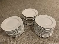 Three stacks of white ceramic dinner plates on carpet, illustrating the lot quantity and condition.