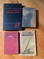 Four vintage cookbooks arranged on wooden floor: Rumford Complete Cookbook, Aunt Sammy's Radio Recipes Revised, Good Housekeeping Institute Cookbook, and Searchlight Recipe Book. Covers show wear and age.