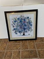 Framed square artwork on floor with visible black tree trunk against white background decorated with blue and silver snowflakes, circular ornaments, and white dots.