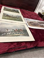 Three art prints laid out on a red fabric surface. Prints depict horse-hunting scenes and horse trading.