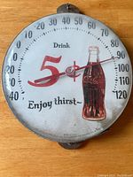 Front of round vintage Coca-Cola thermometer showing branding and temperature dial.