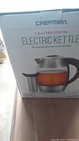 Photo of the Chefman 1.8-liter digital electric kettle packaging showing the kettle with amber liquid inside and removable infuser on the front.