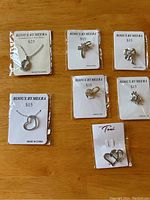 All seven jewelry pieces arranged on a wooden surface, including five pins and two heart pendant necklaces, each mounted on branded display cards.