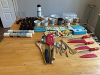 Full view of kitchen items including mugs, glasses, water bottle, utensils, pot holders, napkin rings, ramekins, and kitchen accessories on wooden table.