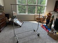 Full view of the metal rolling clothes rack showing height, width and wheels on carpeted floor near a window.