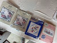 Photo showing three needlepoint pillow kits and one crewel embroidery kit, all still sealed, and two plain white craft towels for stitching projects.
