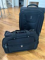 Photo showing two pieces of black Travel Pro luggage, a rollaboard suitcase standing upright and a soft-sided luggage tote in front of it on a hardwood floor.