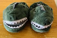 Pair of plush green dinosaur toddler slippers placed side by side on a wooden surface.