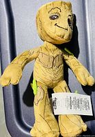 Full view of the Disney Groot plush showing the front of the figure, details including printed bark textures, smiling face, green leaf fabric pieces, and the Disney tag attached near the left arm.