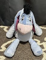 Light blue Eeyore plush toy with pink inner ears, black tuft mane, embroidered eyes and mouth, sitting on carpet.