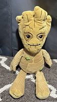 Front view of a Groot plush toy in beige corduroy fabric sitting on a carpet with a plain dark background.