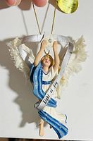 Porcelain angel tree ornament titled 'Light of Joy' by Ashton Drake held by hand for scale, showing detailed blue and white robe, feathered wings, and silver ribbon with text.