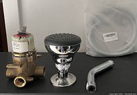 Four plumbing items on a black surface against a white wall: a chrome shower head with attached base, a brass valve, a chrome curved shower arm, and a white coiled hose in plastic packaging.