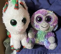 Photo shows two Beanie Boos plush toys, one a white unicorn with striped horn and ears, the other a colorful monkey plush.