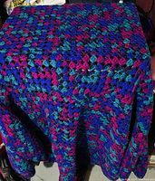 Photo of a crocheted couch throw draped over a surface, showing a multicolored pattern mainly of purple, blue, teal, and pink yarn.