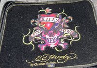 Black fabric car mat with colorful Ed Hardy skull and heart graphic, including text and signature.