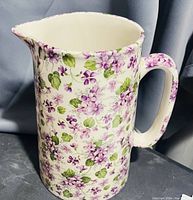 Front and side view of glossy cream-colored ceramic pitcher with purple flowers and green leaves, showing handle and spout.