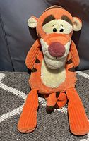Front view of Tigger plush toy with characteristic orange, cream and brown colors; made of corduroy fabric with a pink nose and embroidered eyes, sitting on a patterned carpet.