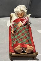 Full view of vintage Mrs. Santa Claus figurine lying in wooden bed covered by red and green plaid blanket with red satin edges, wearing striped red and white top and lace cap.