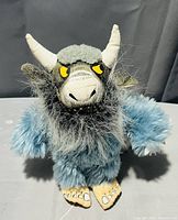 Blue Bernard plush toy with blue fur, grey face, horns, yellow eyes and detailed feet on a flat surface, photographed against a dark background.
