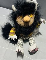 Plush toy of Moishe sitting on a gray surface, showing front view with black fur, yellow eyes, small white horns, striped yellow and black arms, and scaled patterned legs with a pink stain on the foot.