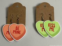 Photo shows two pairs of heart shaped dangle earrings on cardboard holders. The orange pair reads Be Mine in red text and the green pair reads Kiss Me in red text. Both have metal hooks for wearing.