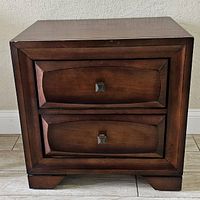 Front view of brown wood nightstand showing two drawers with metal square knobs and a recessed panel design.