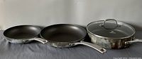 Three Calphalon Classic frying pans, two without lids, one with glass lid, showing front and sides with visible signs of use.
