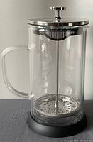 Close-up front view of the cylindrical clear glass French press with handle, stainless steel lid and plunger, sitting on black base.