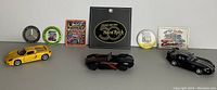 Photo showing three die-cast car models (yellow, black vintage, black modern), two Mercedes emblems, three assorted magnets, and one Hard Rock patch.
