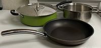 Four cooking pans displayed, including a black non-stick frying pan, a green enameled pan with glass lid, and two stainless steel saucepans with different handle types.