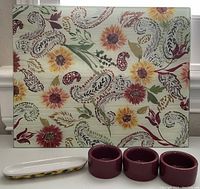 Front view of rectangular glass cutting board with colorful floral and paisley designs and three maroon ceramic sauce holders with small matching oval tray.