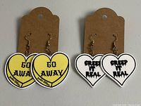 Photo of two pairs of heart-shaped dangle earrings on display cards, one pair yellow with 'Go Away' text, other white with 'Creep It Real' text.