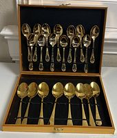 Open case showing 6 large and 12 small gold tone spoons held in place by elastic bands on black cloth interior.