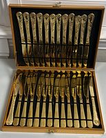 Open gold-tone case showing 12 large knives on top and 12 butter spreaders below, all with embossed decorative handles.