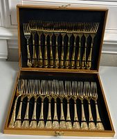 24 gold tone large forks neatly arranged in two rows inside an open black lined gold tone case with a hinged lid.