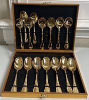 Gold tone large spoons arranged inside a hinged presentation case with a black fabric interior lining and slots for each spoon.